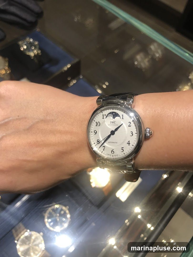 A close-up shot of a person's wrist wearing a silver IWC watch with a white dial. The watch features Arabic numerals, a moon phase complication at the 12 o'clock position, and the text "AUTOMATIC" below the center. The background shows a blurred display case with other watches and jewelry, suggesting a retail environment.