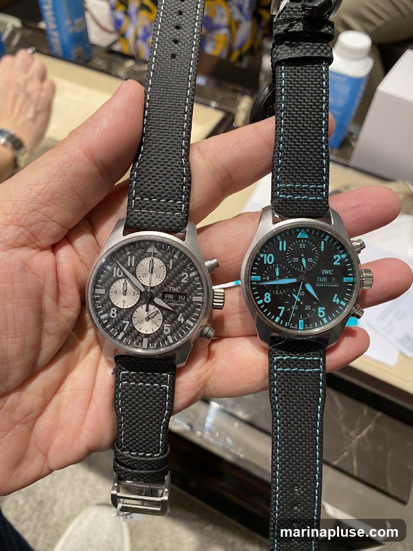 A close-up shot of two IWC watches being held by a person. The watch on the left has a carbon fiber dial with three subdials and displays "FRI 10". The watch on the right has a black dial with luminous blue markers and hands, and displays "TUE 7". Both watches have black fabric straps with contrasting stitching. The background appears to be a retail environment with blurred figures and displays.