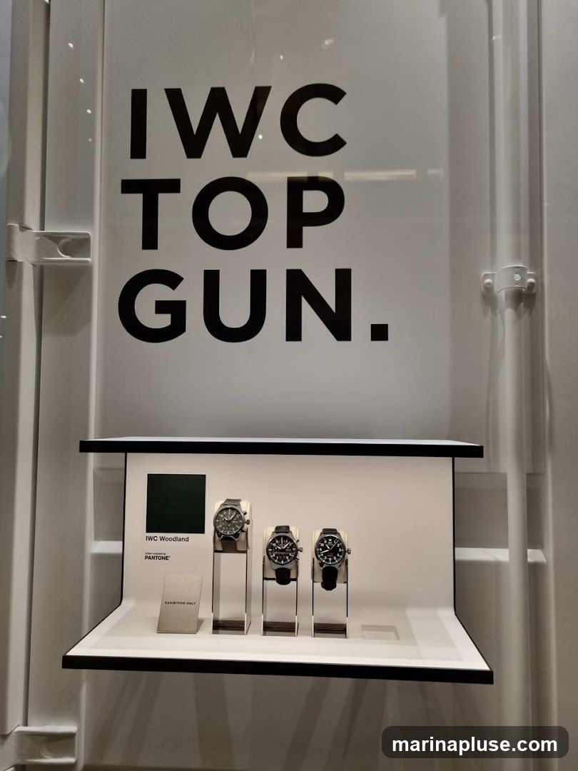 A display featuring three IWC Top Gun watches. The watches are presented on stands within a white display case. Above the watches, large black text reads "IWC TOP GUN.". To the left of the watches, a dark green square is labeled "IWC Woodland" and "Color created by PANTONE". A small sign reads "EXHIBITION ONLY".