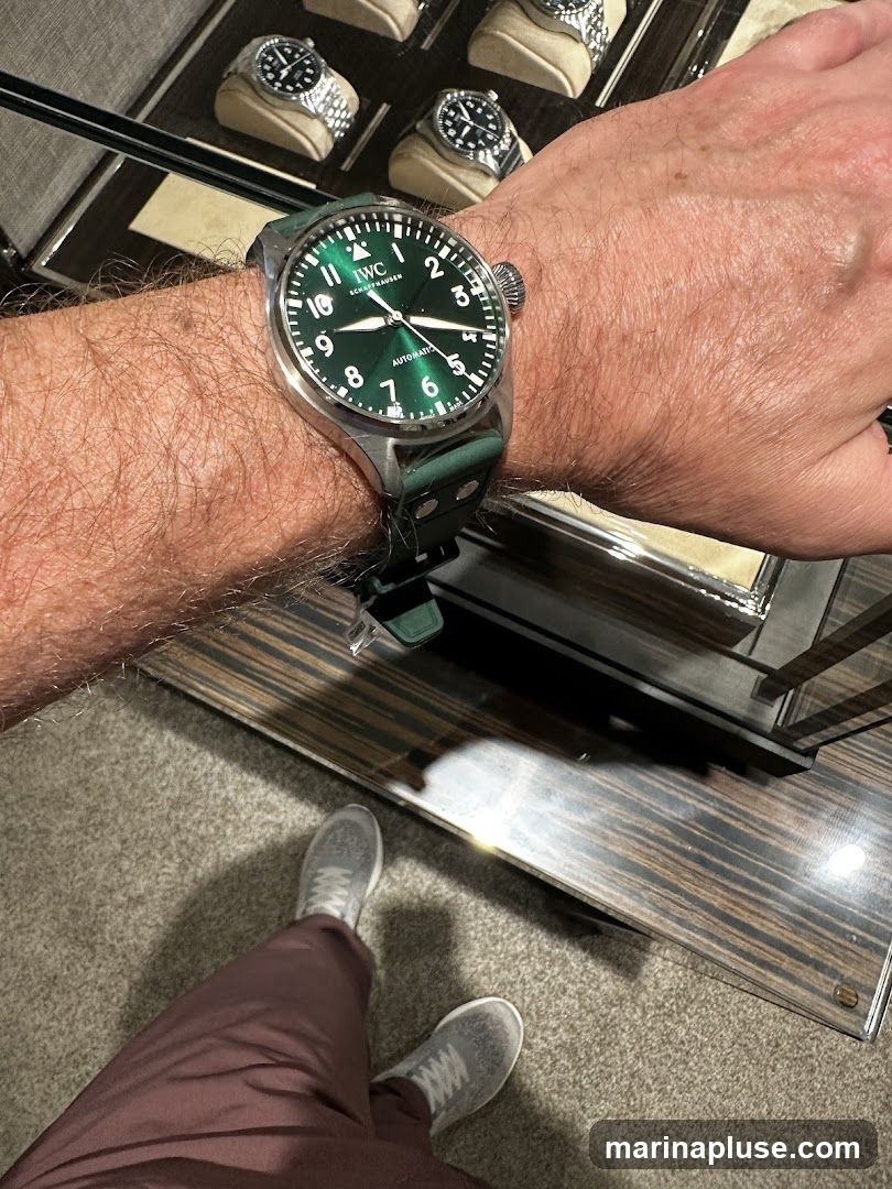 A person's arm wearing a green dial IWC Schaffhausen watch with a dark green strap. In the background, several watches are displayed in a glass case. The person is wearing maroon pants and grey sneakers. The flooring appears to be carpet.