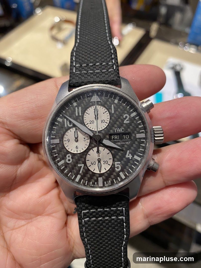 A close-up shot of a person holding an IWC chronograph watch with a black carbon fiber dial and a black fabric strap. The watch face shows the time, day (FRI 10), and date. The background is blurred, showing display cases that suggest a retail environment, possibly a jewelry or watch store.