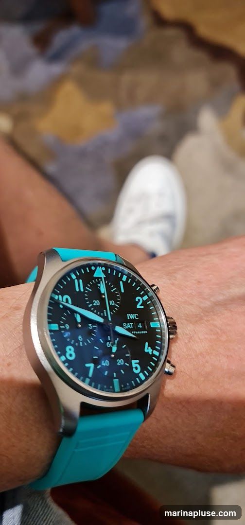 A close-up shot of a person's wrist wearing a watch with a black face and bright blue luminous hands and hour markers. The watch has a silver casing and a bright teal strap. The watch face shows the time and day as Saturday the 4th. In the blurred background, a white sneaker is visible.