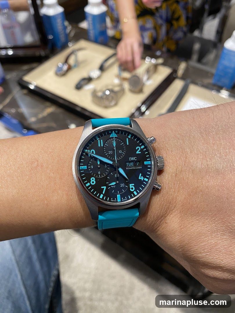 A close-up shot of a person's wrist wearing a watch with a black dial and a bright teal strap. The watch face displays the IWC Schaffhausen logo, day, date, and chronograph sub-dials. In the blurred background, several other watches are displayed on trays, and people are visible.