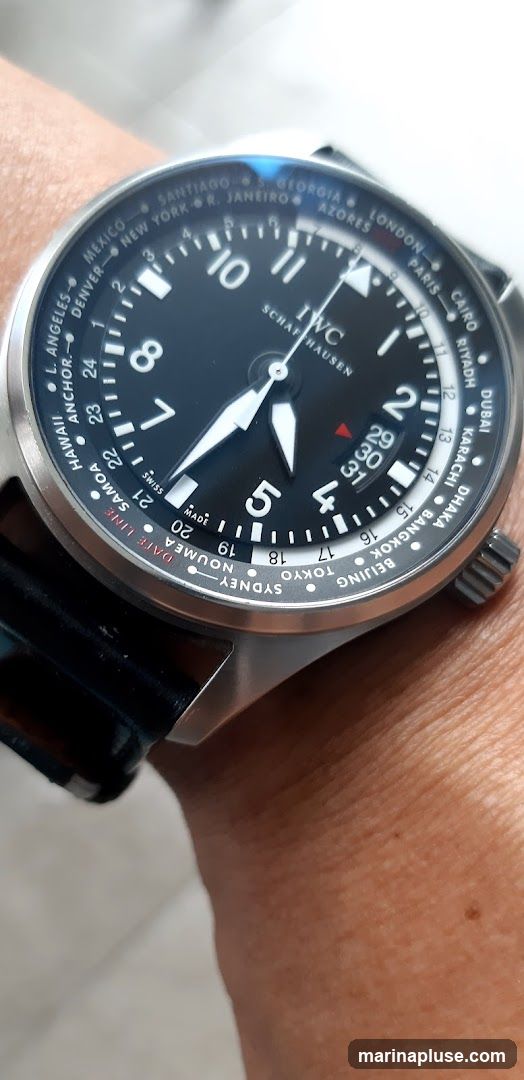 A close-up shot of a man's wrist wearing a black dial IWC Schaffhausen watch with a world timer complication. The watch features city names around the bezel indicating different time zones. The hands point to approximately 7:05. The date window shows the 29th.