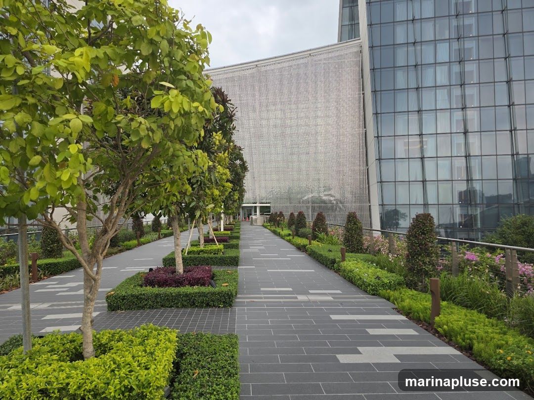 A paved walkway lined with trees and manicured bushes leads towards the entrance of a modern building. The building features a glass facade on one side and a textured, perforated metal facade on the other. The walkway is bordered by lush greenery and flowering plants.