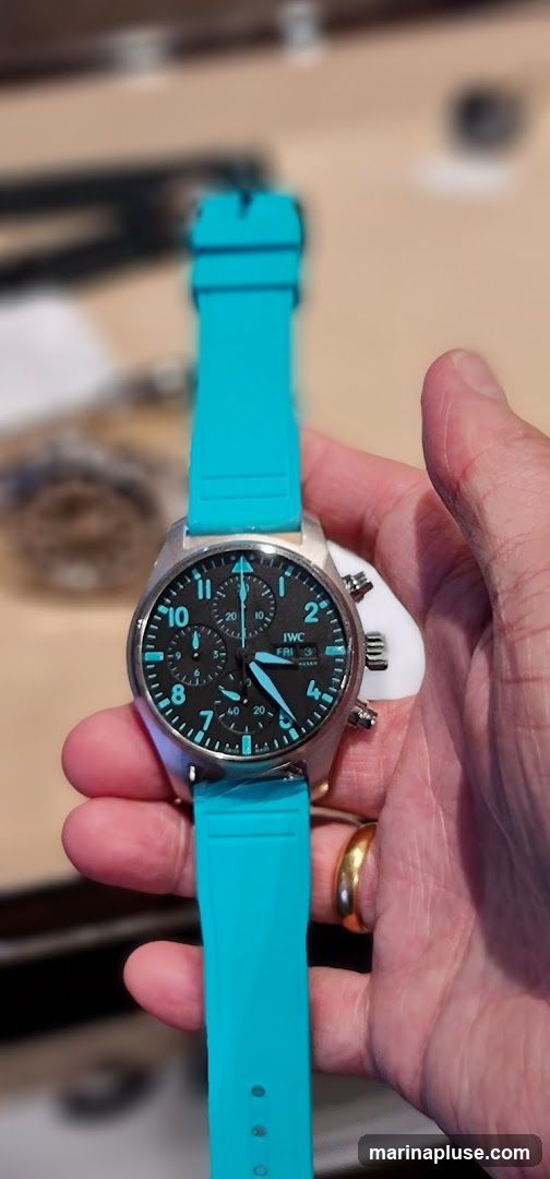 A person's hand holds an IWC Pilot's Watch Chronograph with a bright teal strap. The watch has a black dial with luminous markers and hands, and three subdials. The date window shows "FRI 3". The background is blurred, showing what appears to be a retail environment with lights and displays.