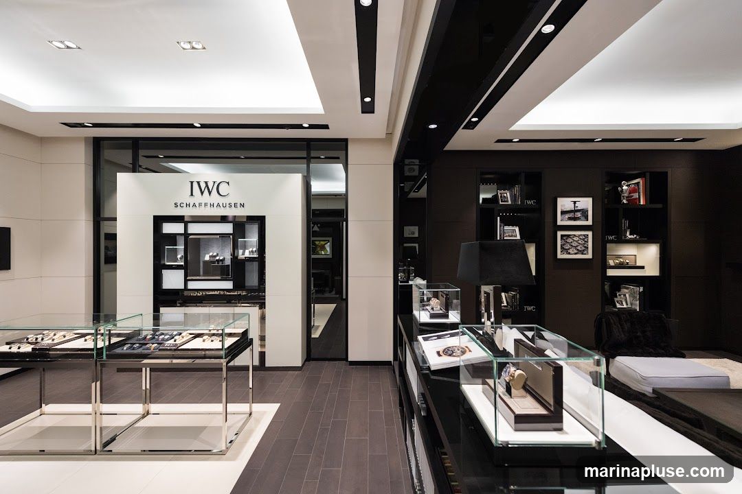 The interior of a luxury watch store, featuring display cases filled with watches. The IWC Schaffhausen logo is prominently displayed on a white pillar. The store has a modern and elegant design with dark wood shelving and ambient lighting.