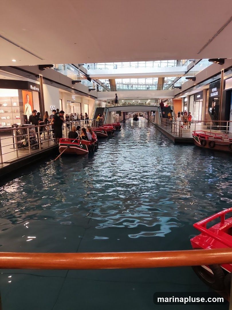 An indoor canal with several gondola-style boats carrying passengers. The canal is surrounded by shops and walkways on multiple levels. The architecture suggests a large shopping mall or entertainment complex.
