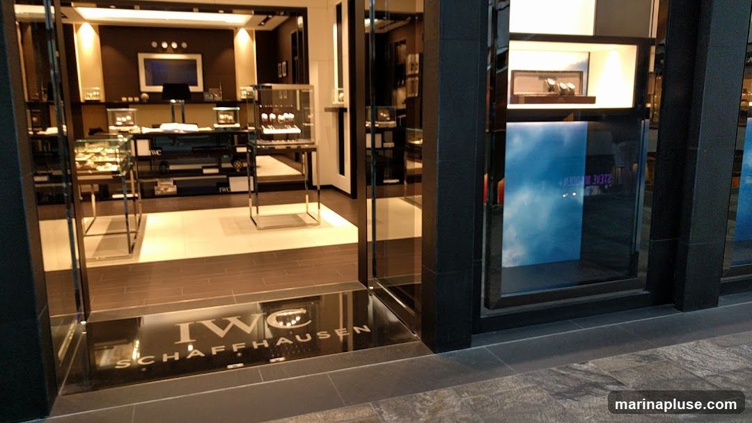 The entrance to a luxury watch store, with display cases visible inside. A doormat with the IWC Schaffhausen logo is placed at the entrance. The store has a modern and sleek design with dark framing and glass windows.