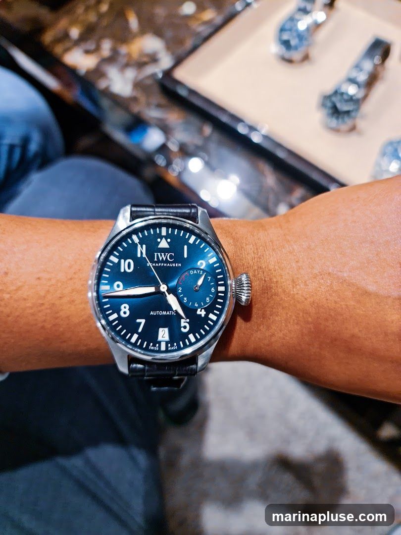 A close-up shot of a person's wrist wearing a blue-faced IWC Pilot's watch with a black leather strap. The watch face displays Arabic numerals, a date window, and a power reserve indicator. In the blurred background, other watches are visible on display.