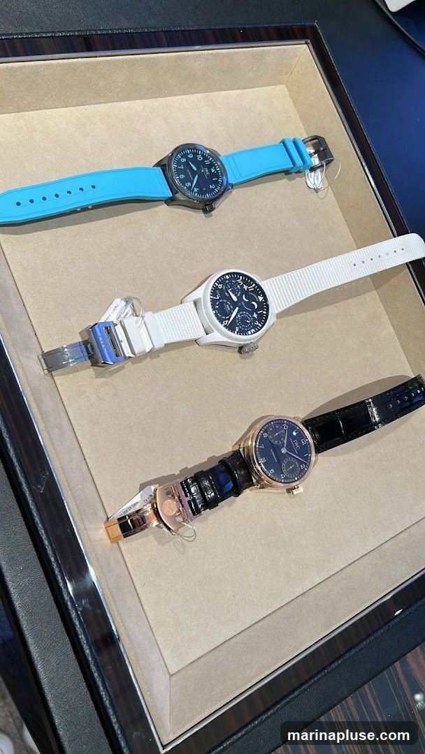A close-up view of three luxury watches, each with a different colored strap, laid out in a display case. The top watch has a bright blue strap. The middle watch has a white strap and a more complex dial with moon phase indication. The bottom watch has a black leather strap and a rose gold case. All watches have visible branding on their dials.