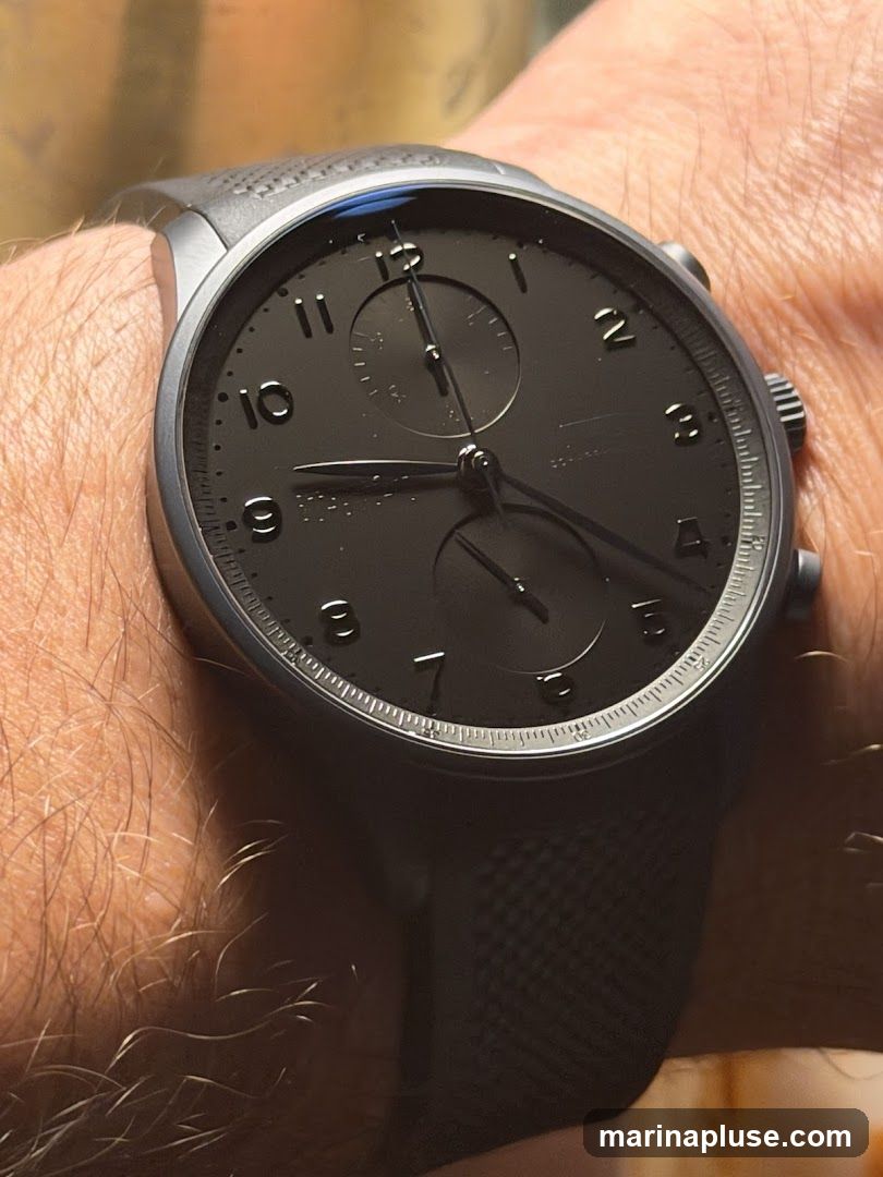 A close-up shot of a dark-faced watch with a black band worn on a hairy wrist. The watch has Arabic numerals for hours and two sub-dials.