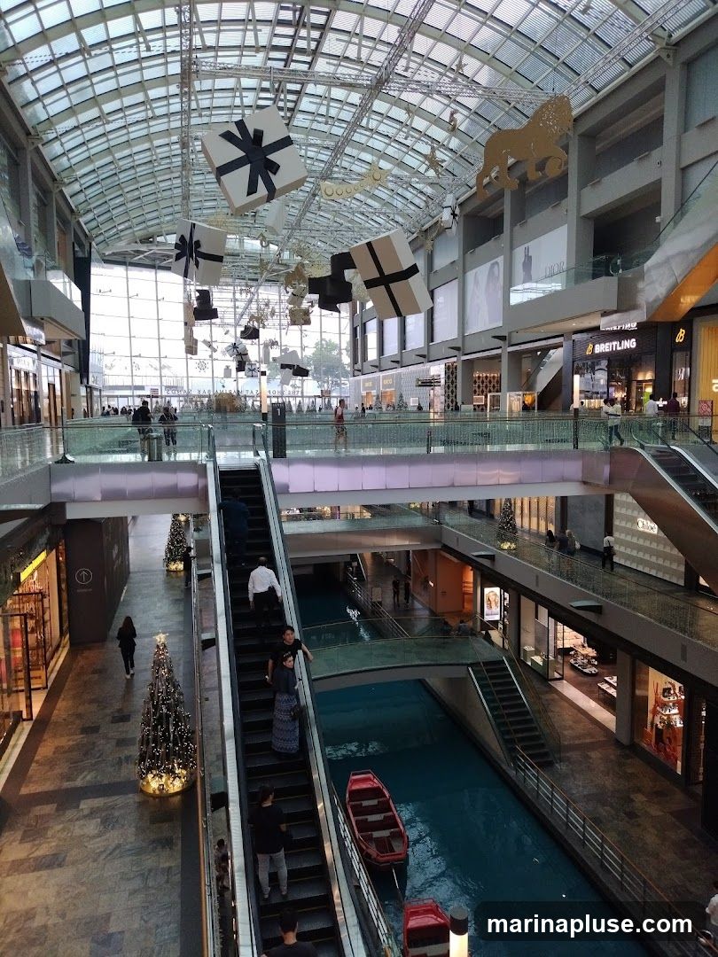 An interior view of a multi-level shopping mall featuring a canal with boats, escalators, and various shops. Large gift boxes and a lion decoration are suspended from the ceiling, suggesting a festive or promotional event. People are seen walking and using the escalators. Storefronts for brands like Dior and Breitling are visible.