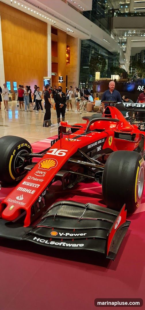 A red Formula 1 race car is on display indoors, with various sponsors' logos visible on its body. The car is positioned on a red surface. In the background, people are walking around in a modern building with wooden paneling and glass structures.