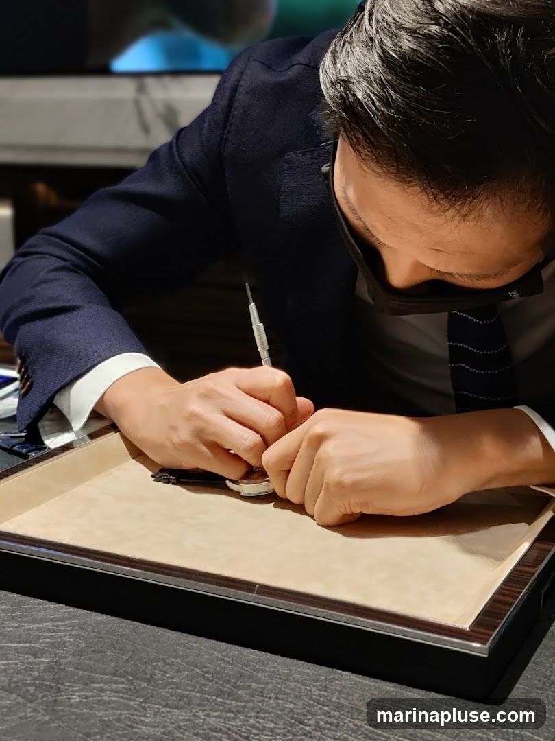 A person wearing a dark suit, white shirt, and tie, with a black face mask, is focused on repairing a watch. They are using a small tool to work on the watch, which is placed on a light brown tray. The background is blurred, suggesting an indoor setting, possibly a workshop or retail store.