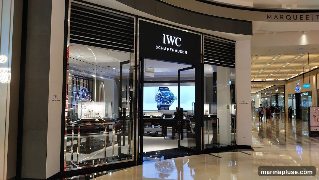 The exterior of an IWC Schaffhausen watch store in a mall. The storefront features large glass windows and doors, with the IWC Schaffhausen logo prominently displayed above the entrance. Inside, display cases showcase watches, and a large screen shows an advertisement for a watch. The mall's interior, including other storefronts and shoppers, is visible in the background.