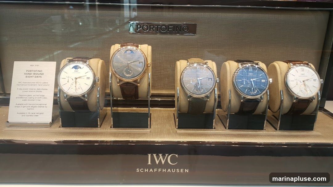 A display case featuring several IWC Portofino watches. The watches have various dial colors and strap materials. A descriptive card for the Portofino Hand-Wound Eight Days watch is visible on the left. The IWC Schaffhausen logo is displayed prominently at the bottom.
