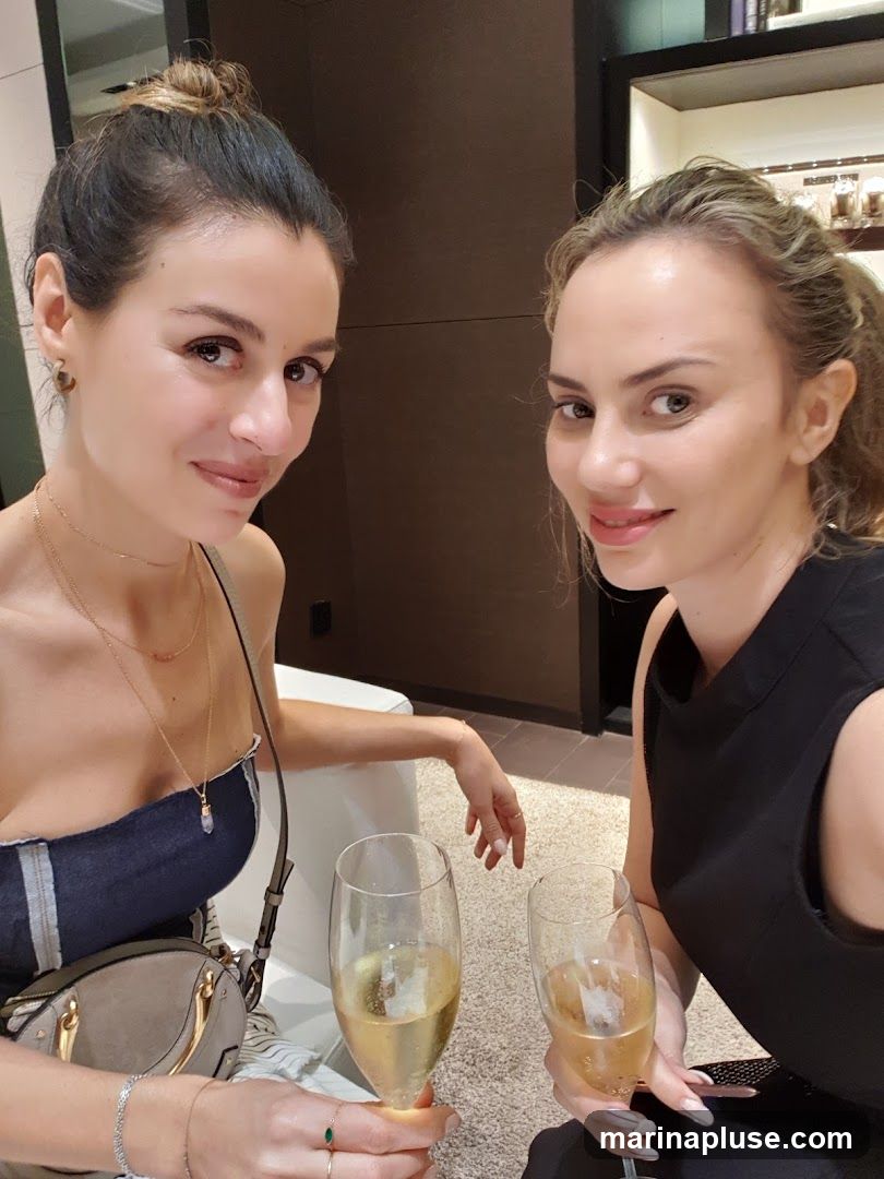 Two women are smiling and holding glasses of champagne. They appear to be in a well-lit, modern interior, possibly a lounge or a high-end store, with shelves displaying items in the background.
