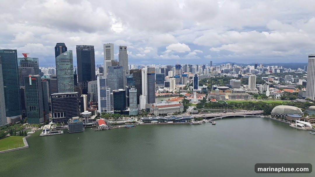 A panoramic view of the Singapore skyline on a cloudy day, showcasing numerous skyscrapers and buildings along the waterfront. The Marina Bay area is visible with its distinctive architecture, including the Esplanade – Theatres on the Bay. The water in the foreground reflects the overcast sky.