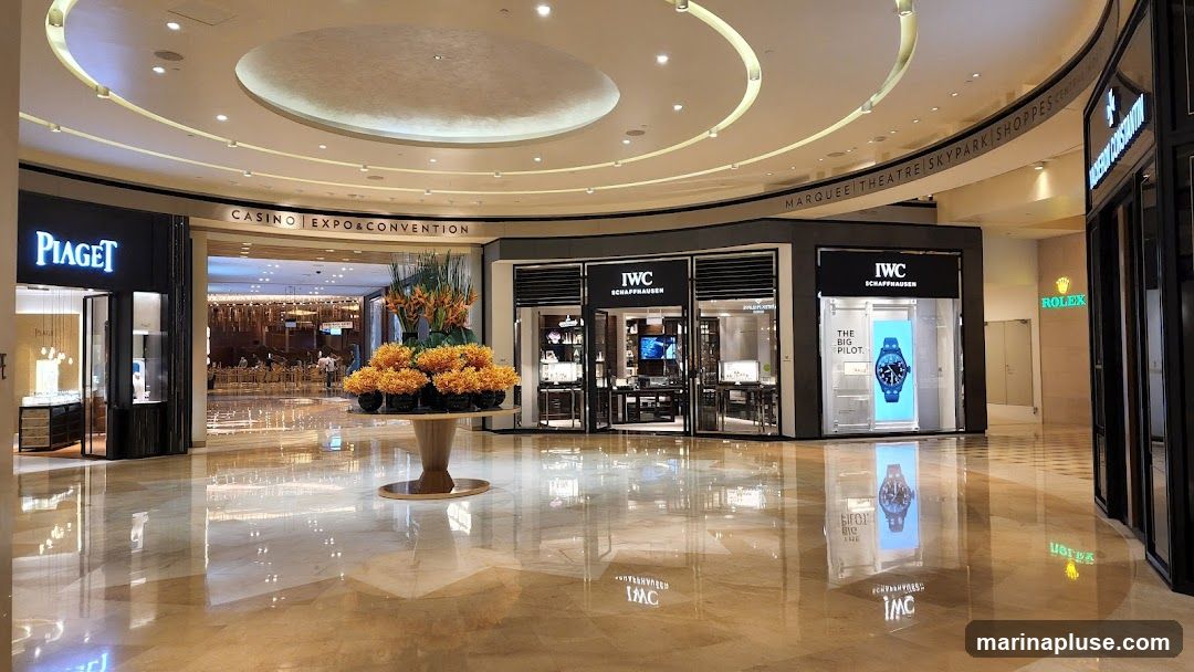 A wide shot of a polished marble floor in a luxury shopping mall. On the left, the entrance to a Piaget store is visible, with its name illuminated. In the center, an IWC Schaffhausen store displays watches in its windows, with a large advertisement for "The Big Pilot" watch. Further to the right, a Rolex store is partially visible. Above the stores, signage indicates "CASINO EXPO & CONVENTION" and "MARQUEE THEATRE SKYPARK SHOPPES". A large floral arrangement sits in the middle of the walkway.