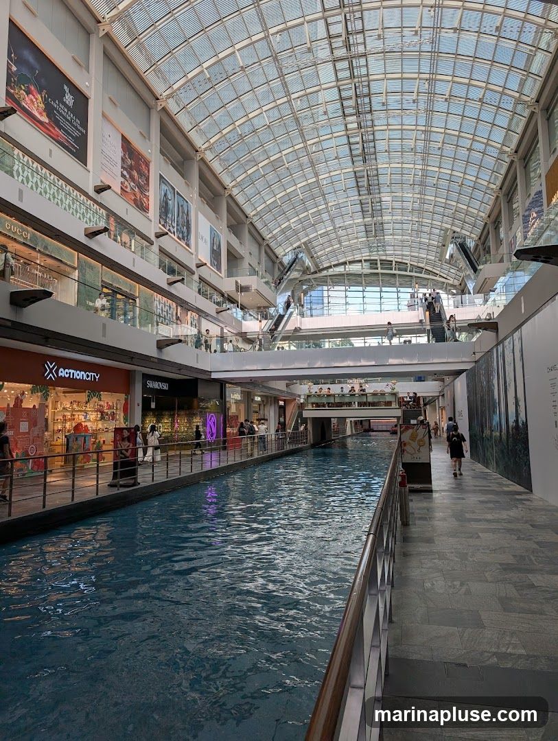 An indoor view of a shopping mall with a canal running through it. Shops are visible on either side of the canal, with escalators and walkways on upper levels. The ceiling is a large glass structure allowing natural light to enter.