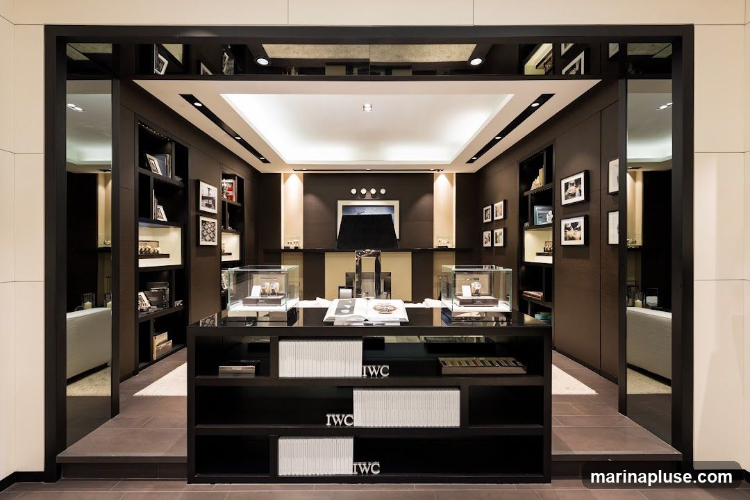The interior of a high-end watch store, featuring dark wood paneling, illuminated display shelves, and a central counter with glass display cases showcasing watches. The IWC logo is visible on the counter and shelves. The store has a modern and sophisticated ambiance.