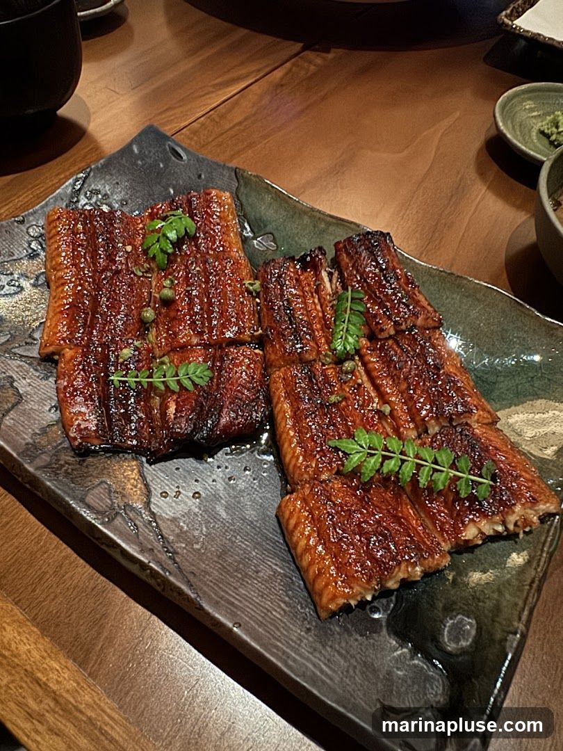 A close-up shot of a dish featuring grilled eel, likely unagi, served on a dark, textured plate. The eel is glazed with a dark, glossy sauce and garnished with fresh green herbs and small green peppercorns. The dish is presented on a wooden table, with hints of other tableware visible in the background.