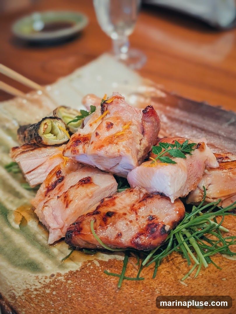 A close-up shot of a plate of grilled chicken pieces, garnished with fresh herbs and orange zest. The chicken appears juicy and slightly charred, served on a textured ceramic plate. In the background, a glass of water and a small dish of sauce are visible on a wooden table.