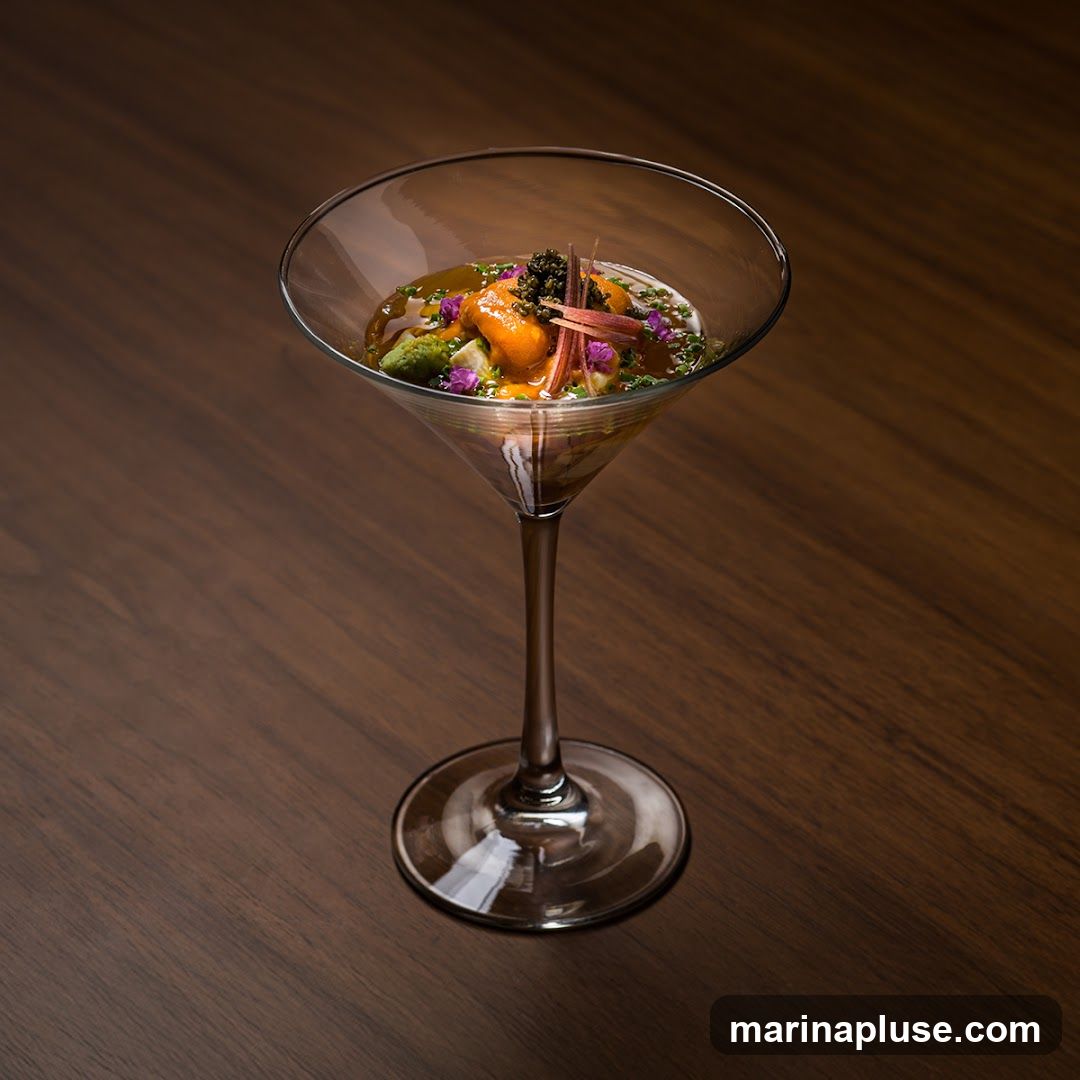 A close-up shot of a delicate appetizer served in a martini glass. The dish features bright orange sea urchin, green vegetables, and black caviar, garnished with thinly sliced ginger and purple edible flowers. The appetizer is presented on a dark wooden surface.
