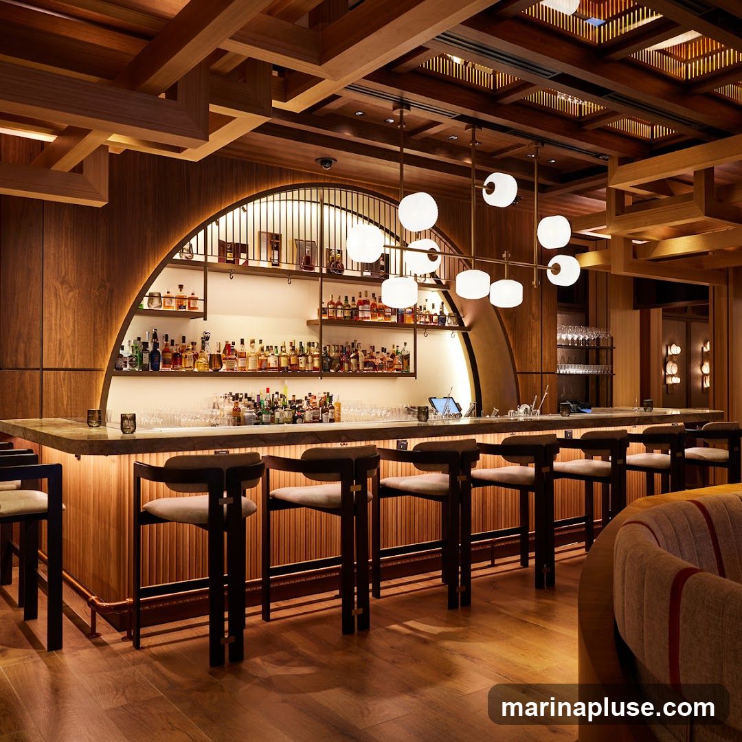 A well-lit bar area with a marble countertop, wooden paneling, and shelves stocked with various bottles of alcohol. Several bar stools are neatly arranged in front of the counter. A modern chandelier hangs from the wooden ceiling.