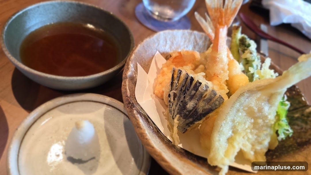 A close-up shot of a tempura platter featuring various fried items including shrimp, eggplant, and vegetables, served with a dipping sauce and grated daikon radish.