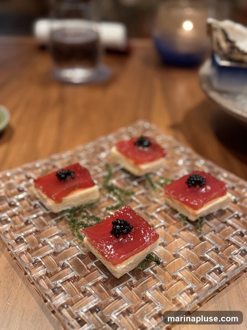Four square appetizers are arranged on a woven glass plate. Each appetizer consists of a light-colored base topped with a red layer and a small mound of black caviar. Green sprigs are placed around the appetizers. The background is blurred, showing a wooden table, a glass with a dark liquid, a lit candle in a blue holder, and an oyster shell on a plate.