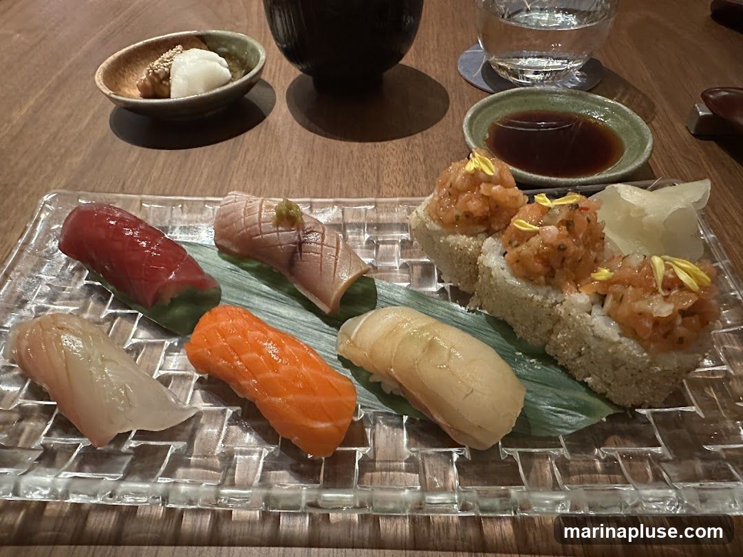 A close-up shot of a sushi platter featuring various types of sushi and a small dish of pickled ginger. The platter includes tuna, salmon, and other fish nigiri, as well as what appear to be spicy tuna rolls. A small bowl with what looks like pickled radish and sesame seeds is also visible, along with a dish of soy sauce and a glass of water.