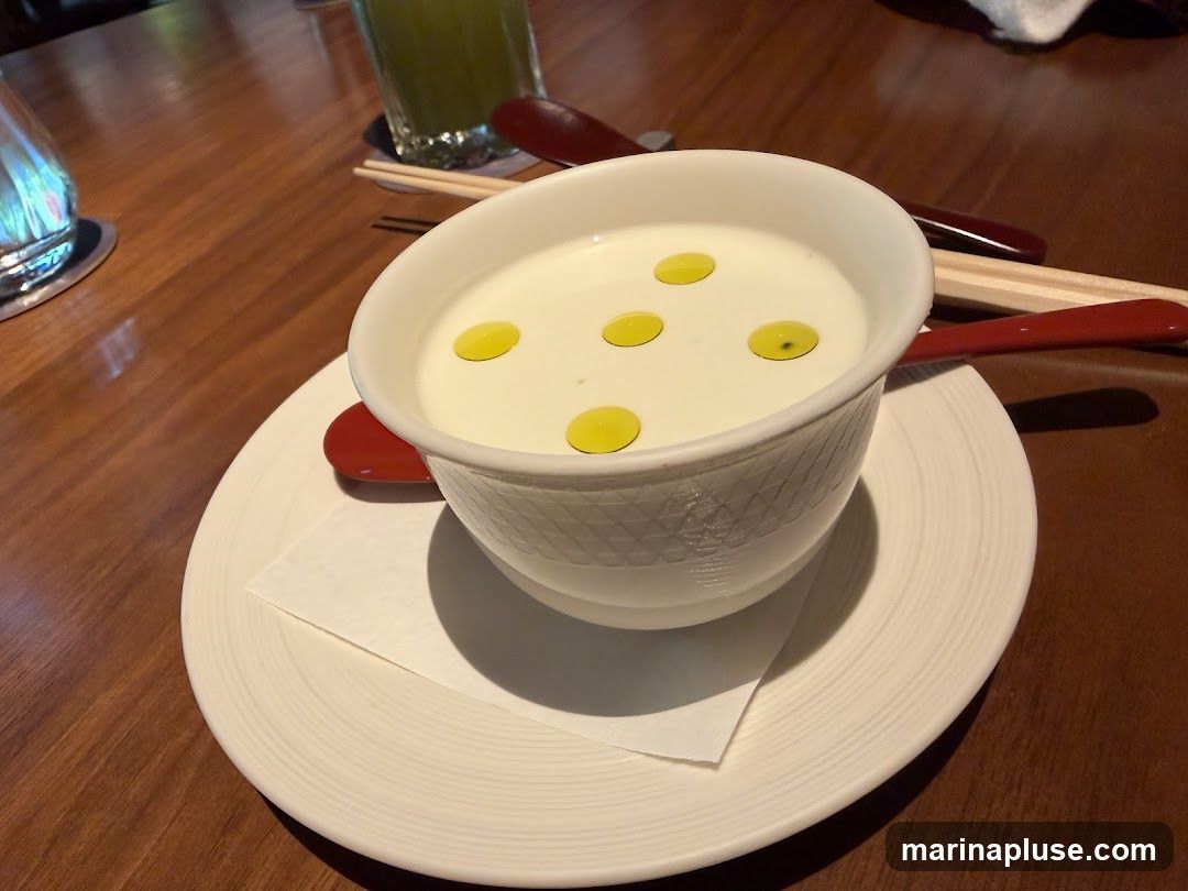 A close-up shot of a white bowl filled with a creamy white soup, garnished with several drops of yellow olive oil. The bowl sits on a white saucer with a textured rim, placed on a wooden table. A red spoon and chopsticks are visible behind the bowl, along with a glass of green liquid in the background.