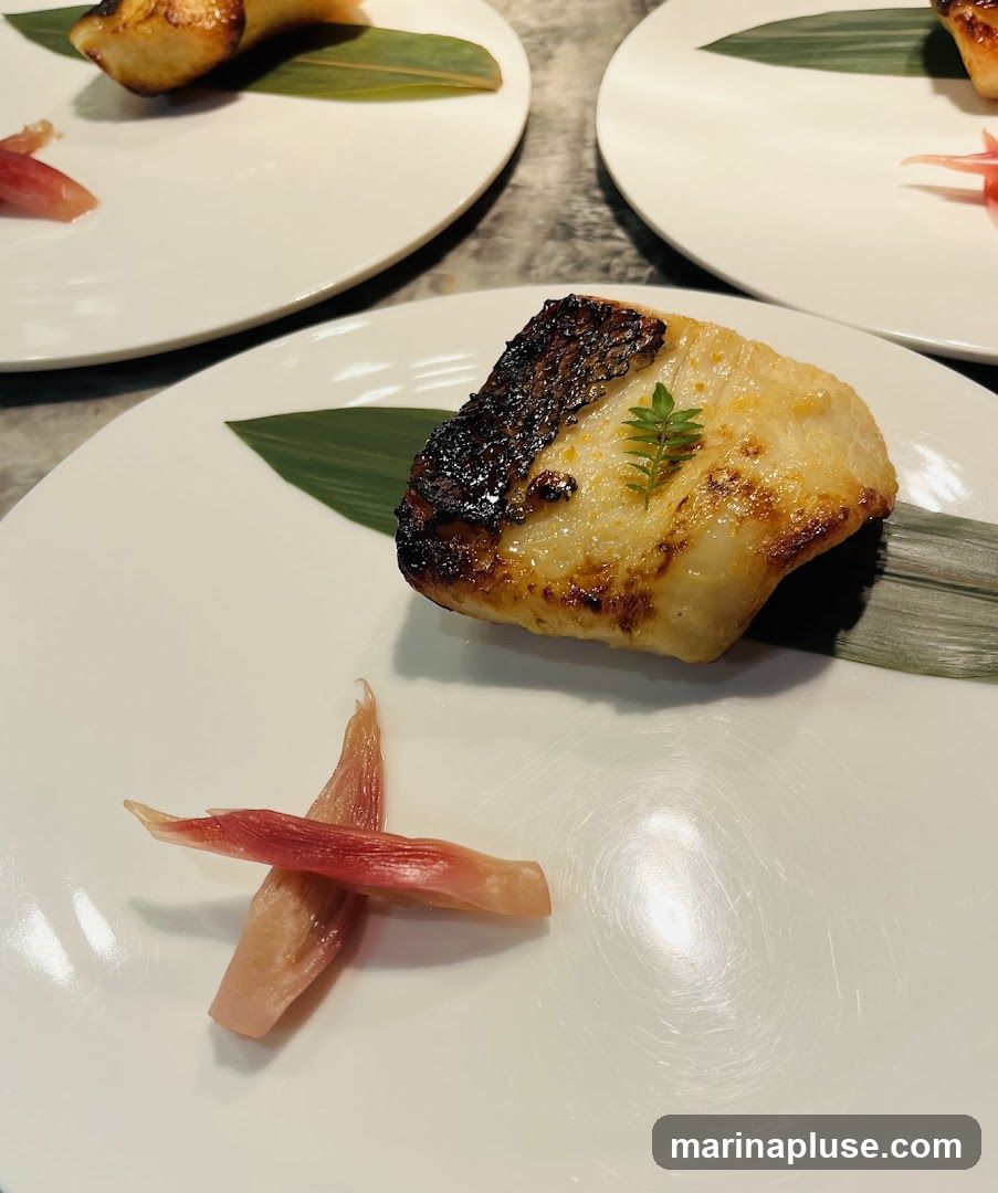 A close-up shot of a piece of grilled fish served on a white plate. The fish is golden brown with some charred edges and is garnished with a small green leaf. A side of pickled ginger, cut into thin strips, is also on the plate. The dish is presented on a textured surface, and other plates with similar food are visible in the background.