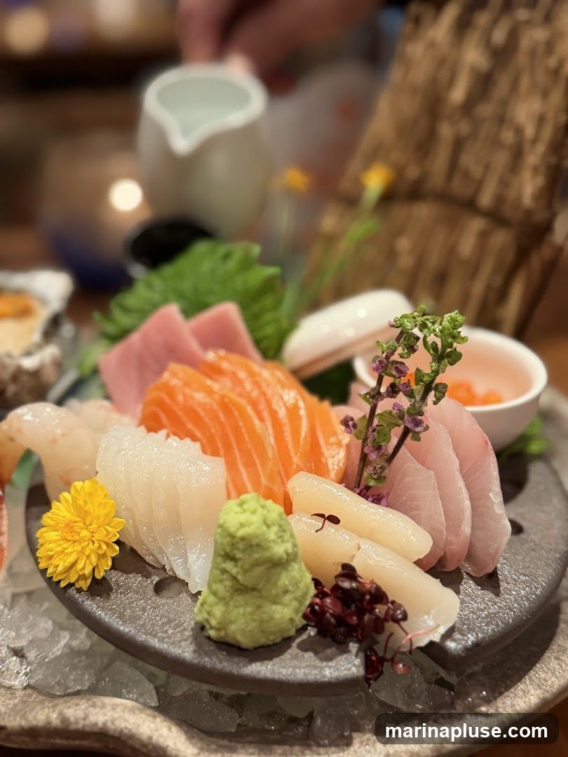 A close-up of a sashimi platter featuring various types of fish, including salmon and tuna, arranged on a dark plate with wasabi, a yellow flower, and microgreens. A small pitcher of sauce is being poured in the background.