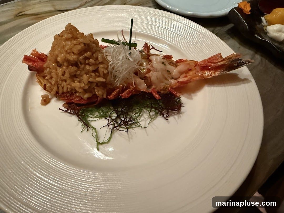A close-up shot of a white plate with a large shrimp served on top of a mound of rice. The shrimp is split lengthwise, revealing its cooked meat. The rice appears to be a savory pilaf or risotto. The dish is garnished with shredded white vegetables and a sprig of green onion. Dark green and red seaweed are placed around the base of the shrimp. In the background, a small dark dish holds what appears to be fruit and a dollop of cream.