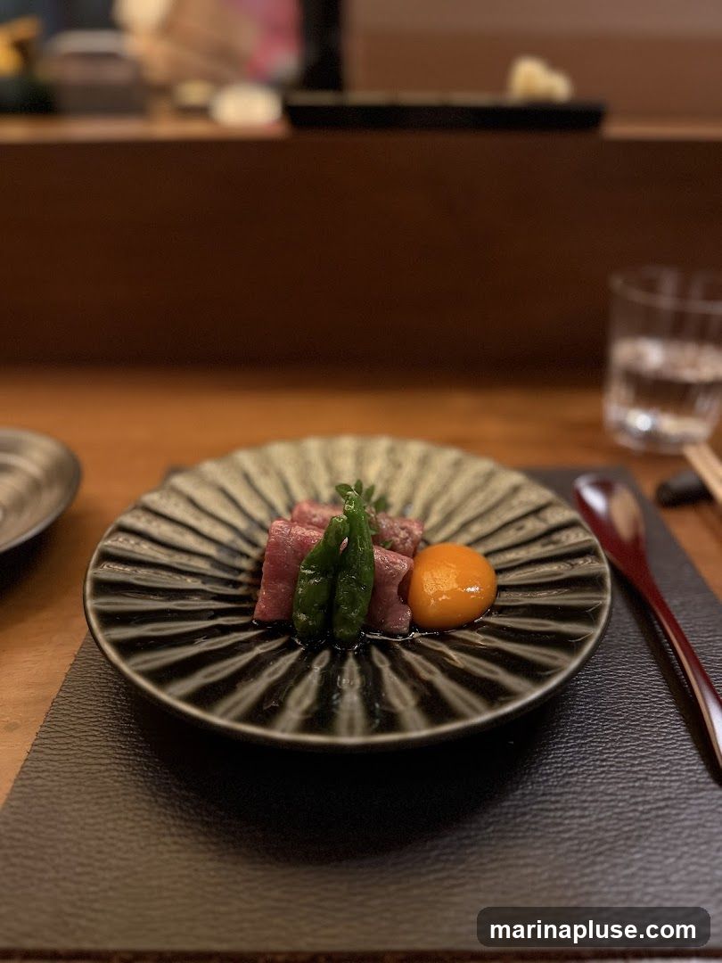 A close-up shot of a small appetizer dish featuring thinly sliced beef, two green vegetables, and a dollop of bright orange sauce, possibly egg yolk. The dish is served on a dark, ridged plate, which rests on a textured placemat. In the background, a glass of water and chopsticks are visible, suggesting a dining setting.