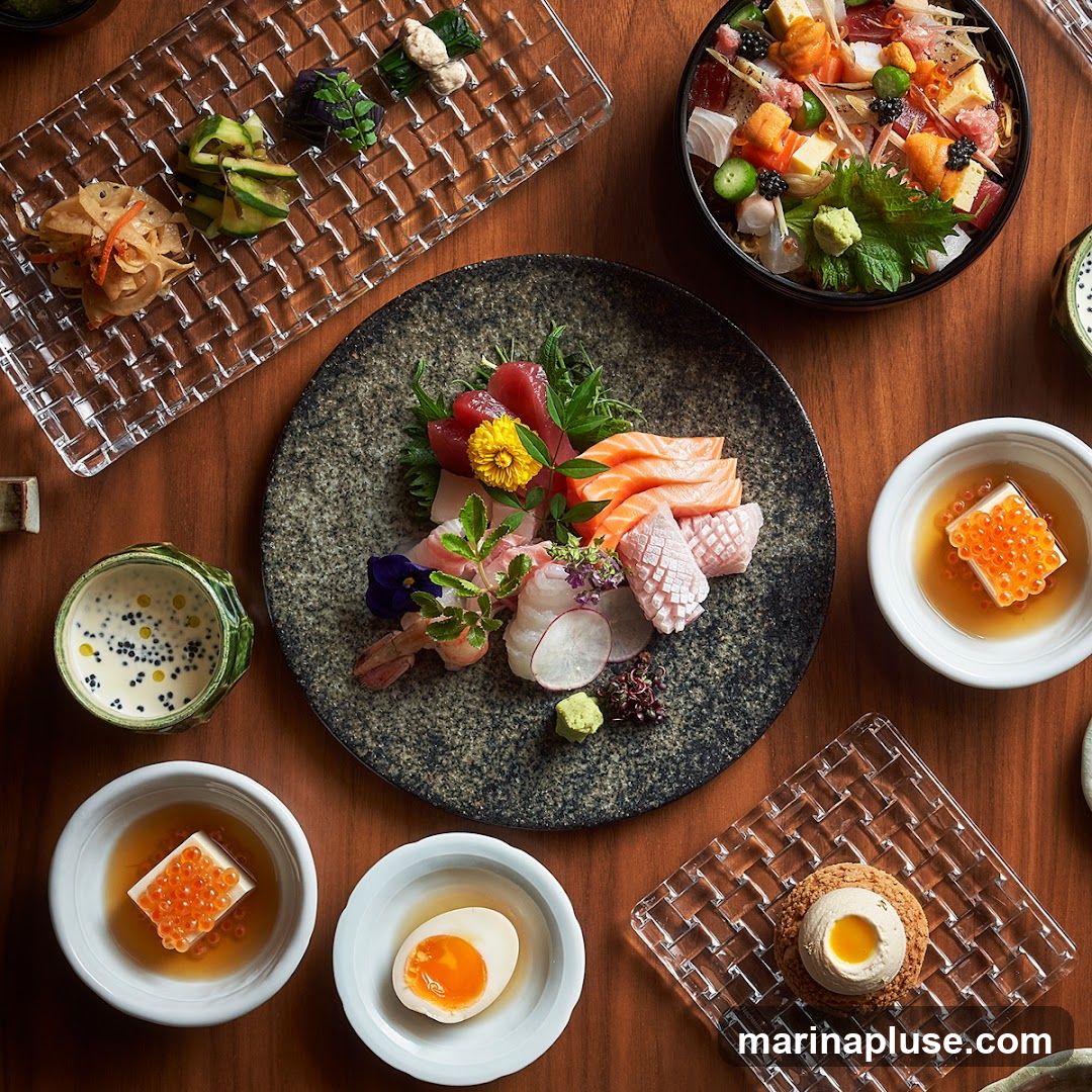 A top-down view of a wooden table laden with various Japanese dishes. In the center, a dark, textured plate holds a vibrant sashimi platter with tuna, salmon, and white fish, garnished with edible flowers and microgreens. To the upper right, a bowl overflows with chirashi sushi, a colorful mix of seafood, egg, and vegetables. Other dishes include a glass tray with pickled vegetables, small bowls of tofu with salmon roe, a soft-boiled egg, and a delicate dessert pastry.
