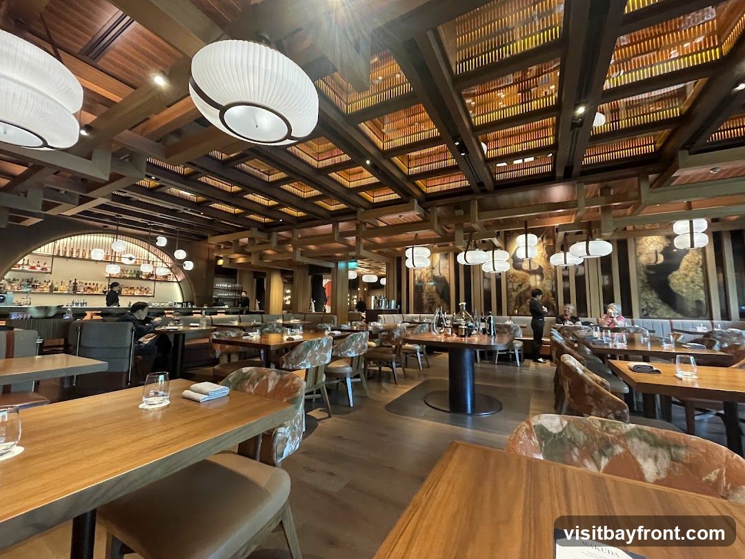 The interior of a modern restaurant with wooden accents, elegant lighting fixtures, and well-set tables. The dining area features a bar in the background, with patrons seated at tables and the bar. The decor includes artistic wall hangings and a slatted wooden ceiling.
