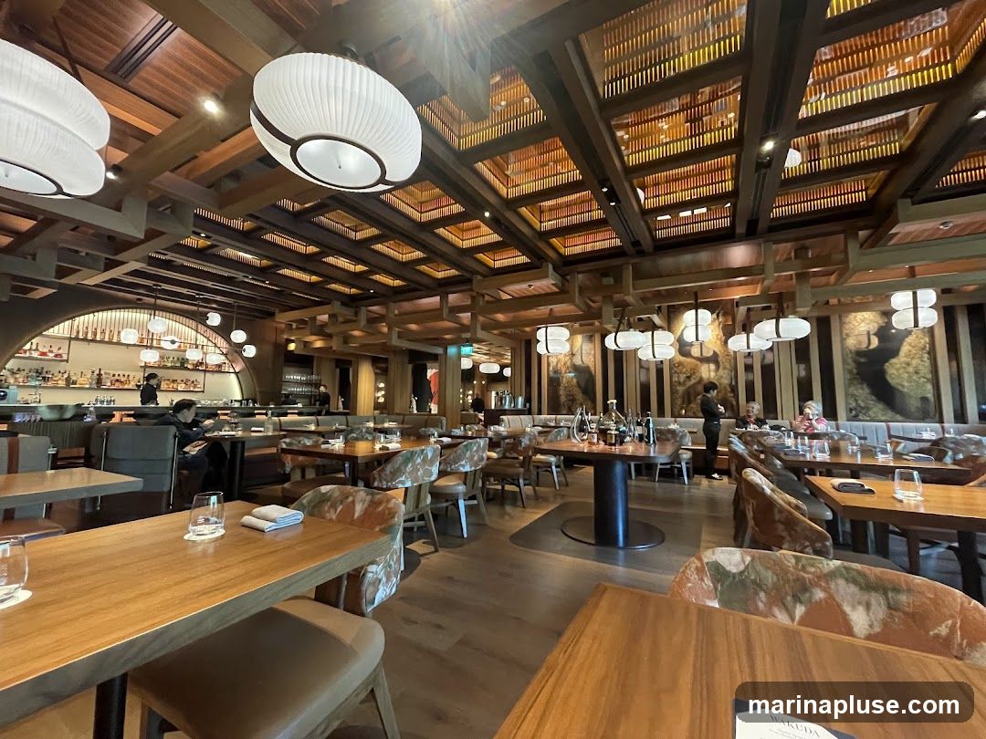 The interior of a modern restaurant with wooden accents, elegant lighting fixtures, and well-set tables. The dining area features a bar in the background, with patrons seated at tables and the bar. The decor includes artistic wall hangings and a slatted wooden ceiling.