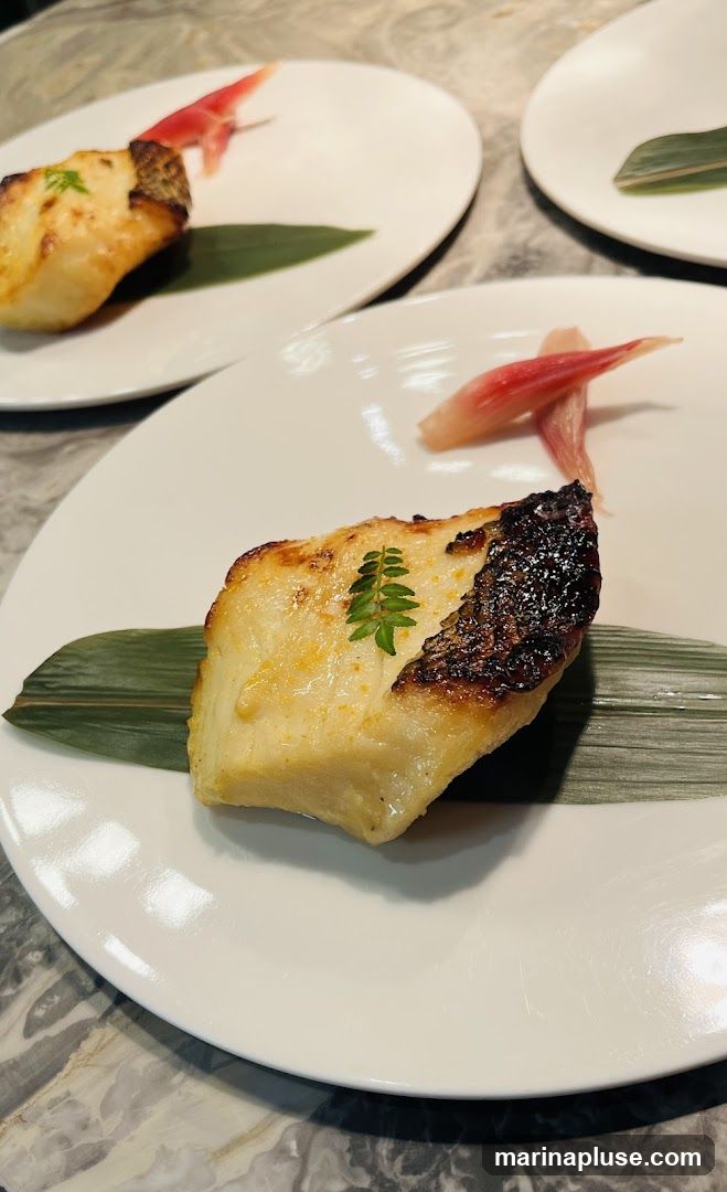 A close-up shot of a piece of seared fish served on a white plate. The fish has a golden-brown crust and is garnished with a small green leaf. A green leaf also serves as a base for the fish. Two pieces of pickled ginger are placed next to the fish. The plate is set on a marble-patterned surface. Multiple plates are visible in the background, suggesting a restaurant setting.