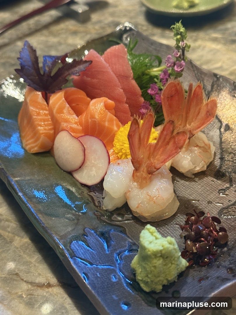 A close-up shot of a sashimi platter featuring slices of salmon and tuna, cooked shrimp, radish slices, wasabi, and garnishes.