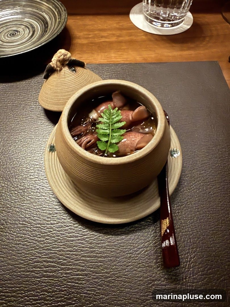 A close-up shot of a small, lidded ceramic pot containing a stewed squid dish. The pot is light brown with ribbed detailing and sits on a matching saucer. The lid, also ribbed, is placed beside the pot and has a small, decorative knot on top. The dish itself contains several small squid in a dark broth, garnished with a sprig of green fern. A dark red spoon with a decorative pattern rests on the saucer. In the background, a textured dark placemat, a wooden table, a patterned bowl, and a glass of water are visible.