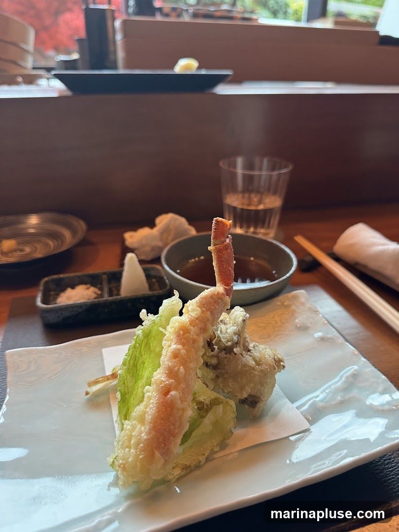 A close-up shot of a tempura dish featuring a piece of shrimp or crab leg, a piece of lettuce, and other fried items. The food is served on a white plate with a small dish of dipping sauce and a side of grated daikon radish. A glass of water and chopsticks are also visible on the table. The background suggests an indoor dining setting with wooden accents.