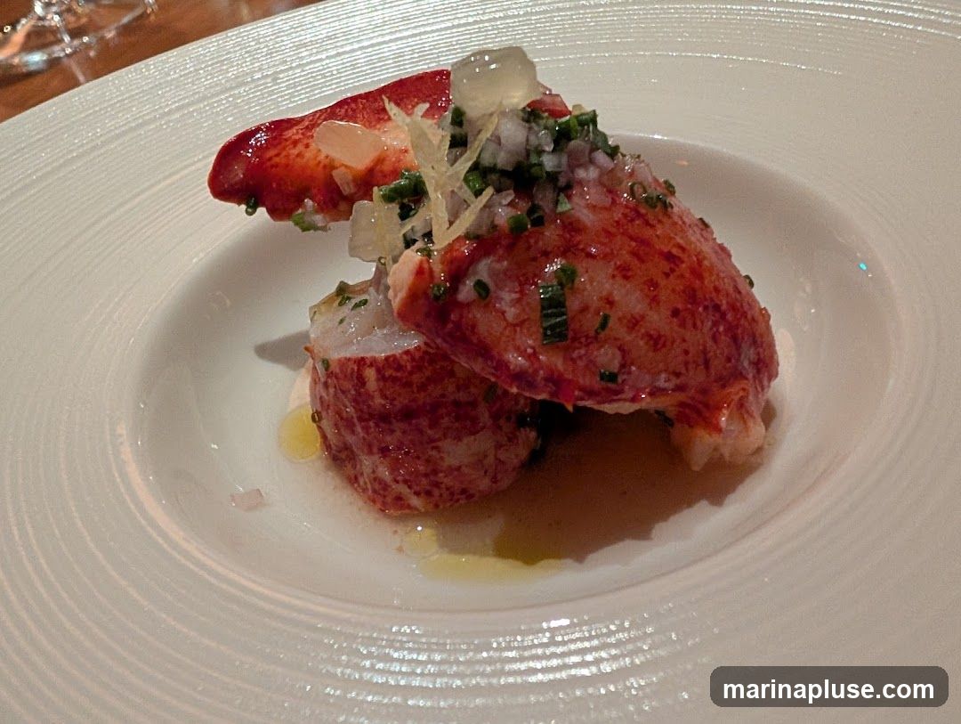A close-up shot of a gourmet appetizer featuring two pieces of cooked lobster claw meat, topped with finely diced onions, chives, and a clear jelly-like substance. The lobster is served on a white, textured plate with a light vinaigrette dressing.