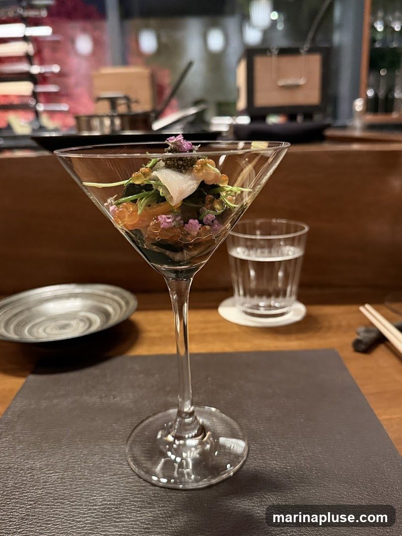 A close-up shot of a delicate appetizer served in a martini glass. The appetizer appears to be layered with various ingredients, including what looks like salmon, roe, and a white fish, topped with caviar and small purple flowers. In the background, a glass of water sits on a coaster, and a dark, textured plate is visible to the left. The setting suggests a fine dining establishment, with blurred elements of the restaurant interior in the background.
