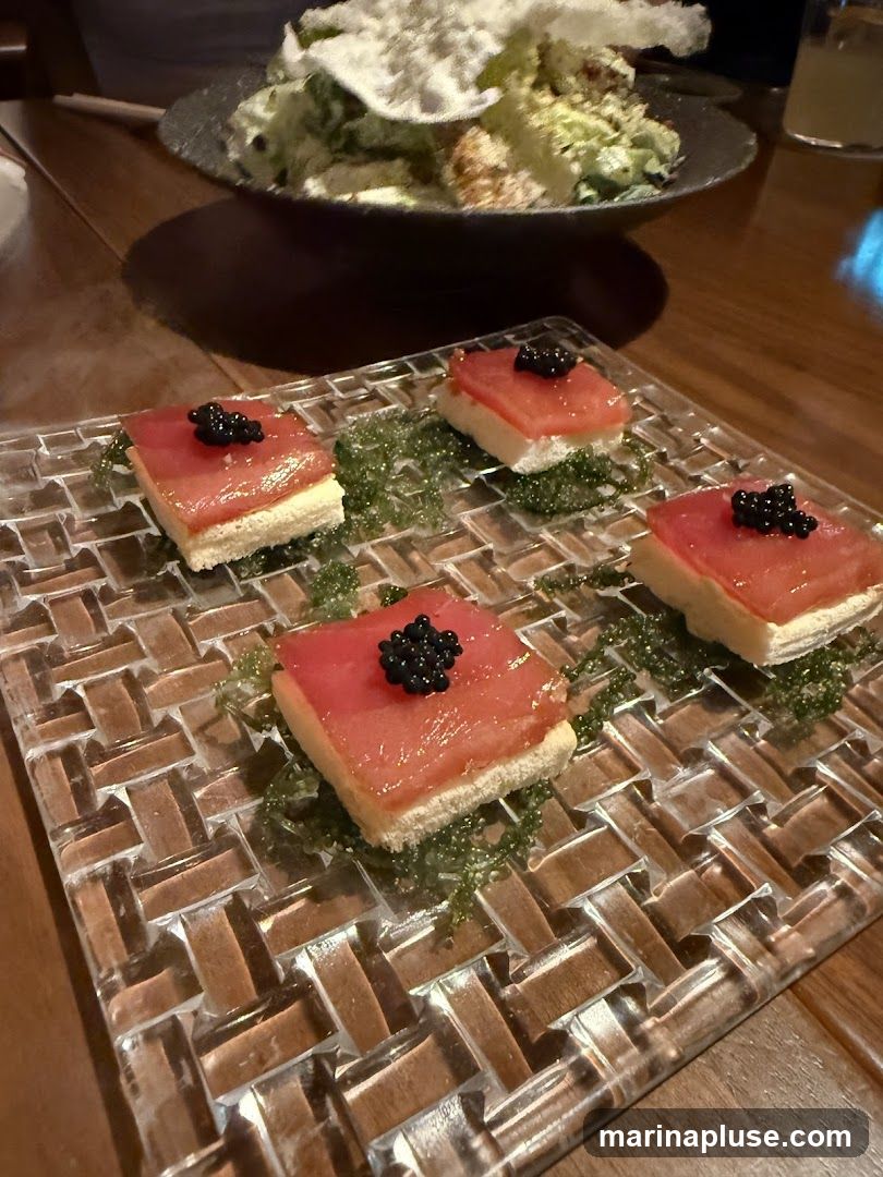 Four square pieces of tuna are served on small white crackers, each topped with black caviar. The bites are arranged on a textured glass plate with a bed of green seaweed. In the background, a salad is visible in a dark bowl.