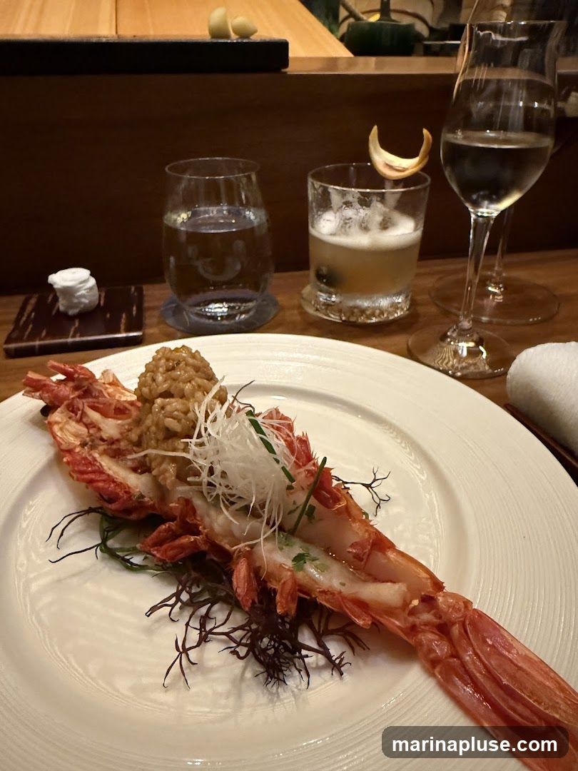 A close-up of a beautifully presented seafood dish, likely a large prawn or lobster tail, served on a white plate with delicate garnishes of shredded white vegetables and microgreens. The dish is accompanied by a small mound of rice or grain. In the background, two glasses of drinks are visible: one with clear liquid and ice, and another a cocktail with foam and a citrus peel garnish. A wine glass with white wine is also present. The setting appears to be a restaurant with warm wooden interiors.