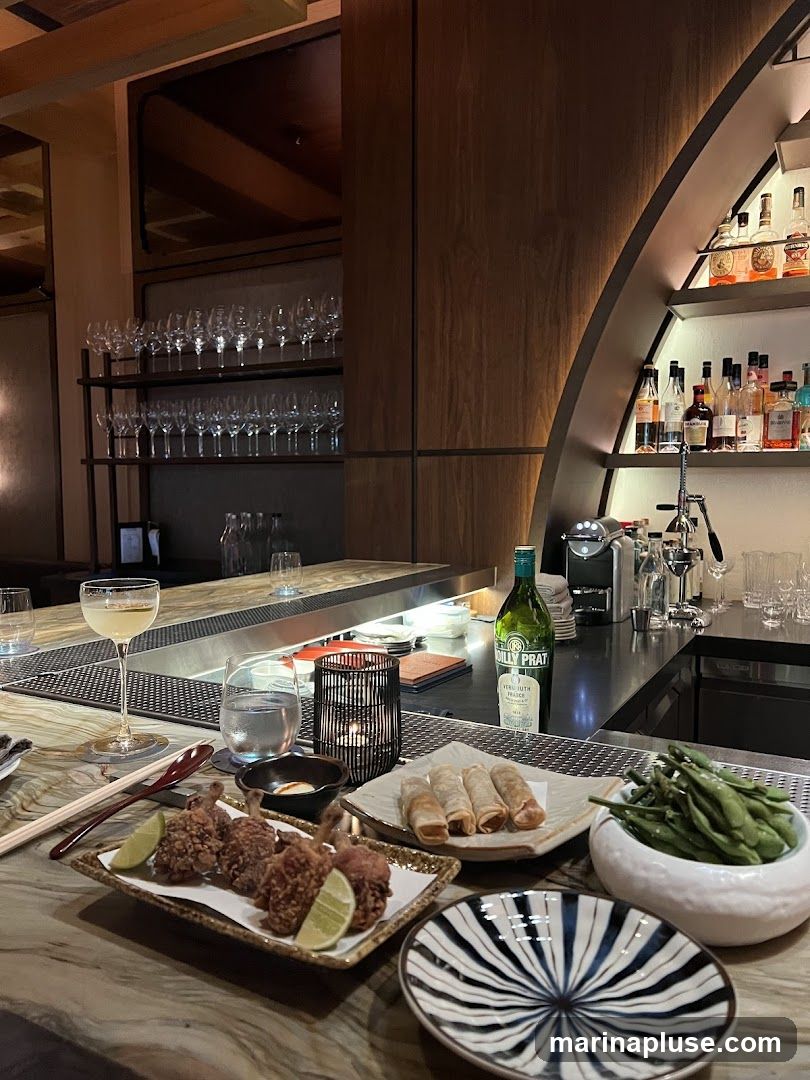 A dimly lit bar area with various appetizers and drinks on the counter. In the foreground, there are plates of fried chicken lollipops, spring rolls, and edamame. A cocktail glass and a bottle of wine are also visible. The background shows shelves stocked with glassware and bottles of liquor.